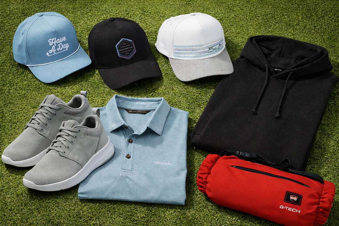 Flat lay of golf apparel and accessories on green turf, including hats, sneakers, a polo shirt, a hoodie, and a G-Tech heated pouch.