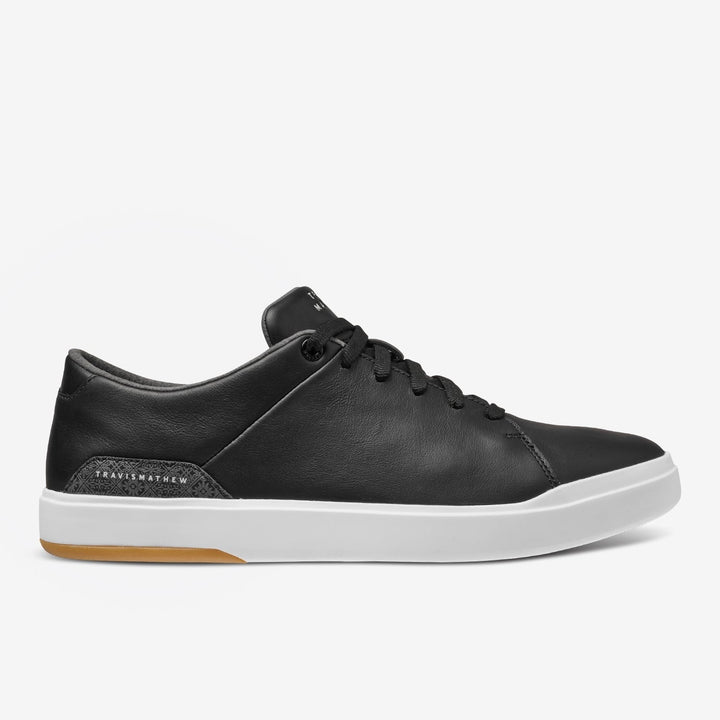 TravisMathew Modern Cup Shoe in black leather with white sole, shown in side profile on a white background.