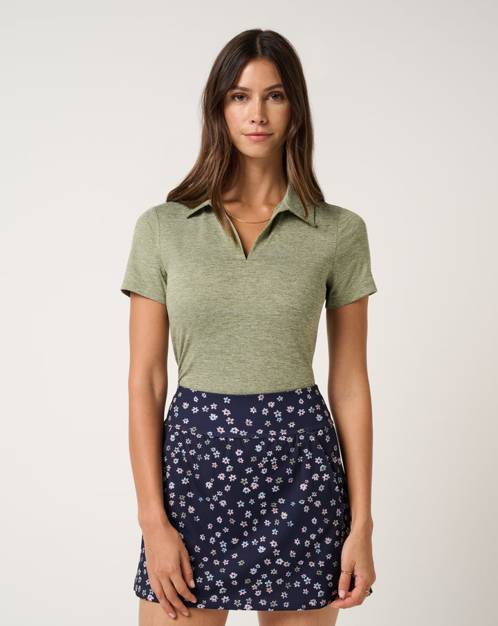 TravisMathew women’s Skyloft Light Polo in heather oil green styled with navy golf skirt.