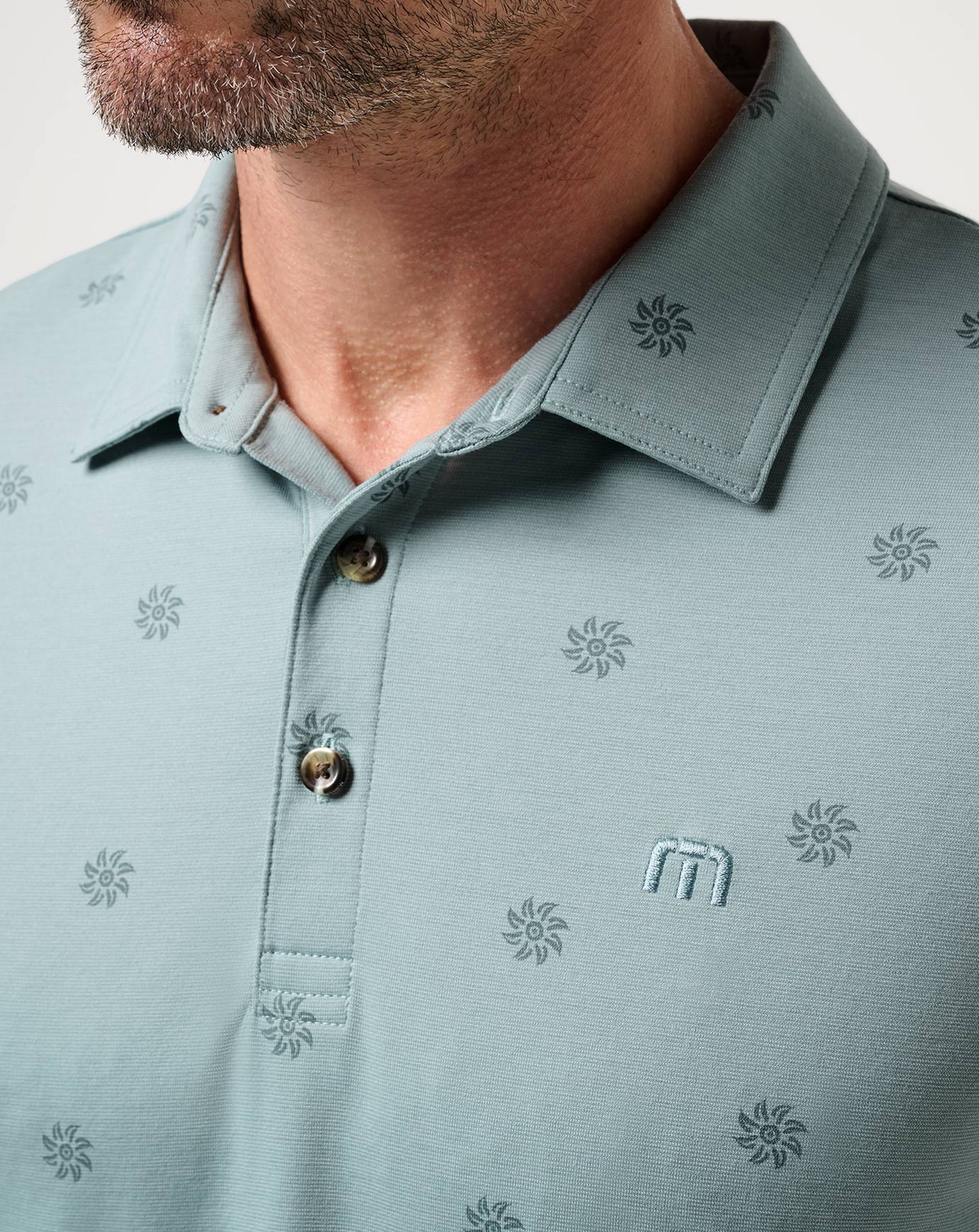 Close-up of the TravisMathew Sol Polo collar, button placket, embroidered logo, and subtle sunburst pattern