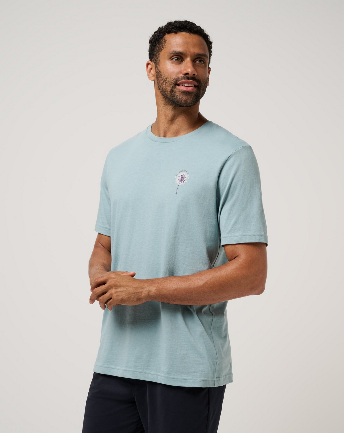 Model wearing the TravisMathew Hold My Calls Tee in light blue with small palm chest graphic, angled