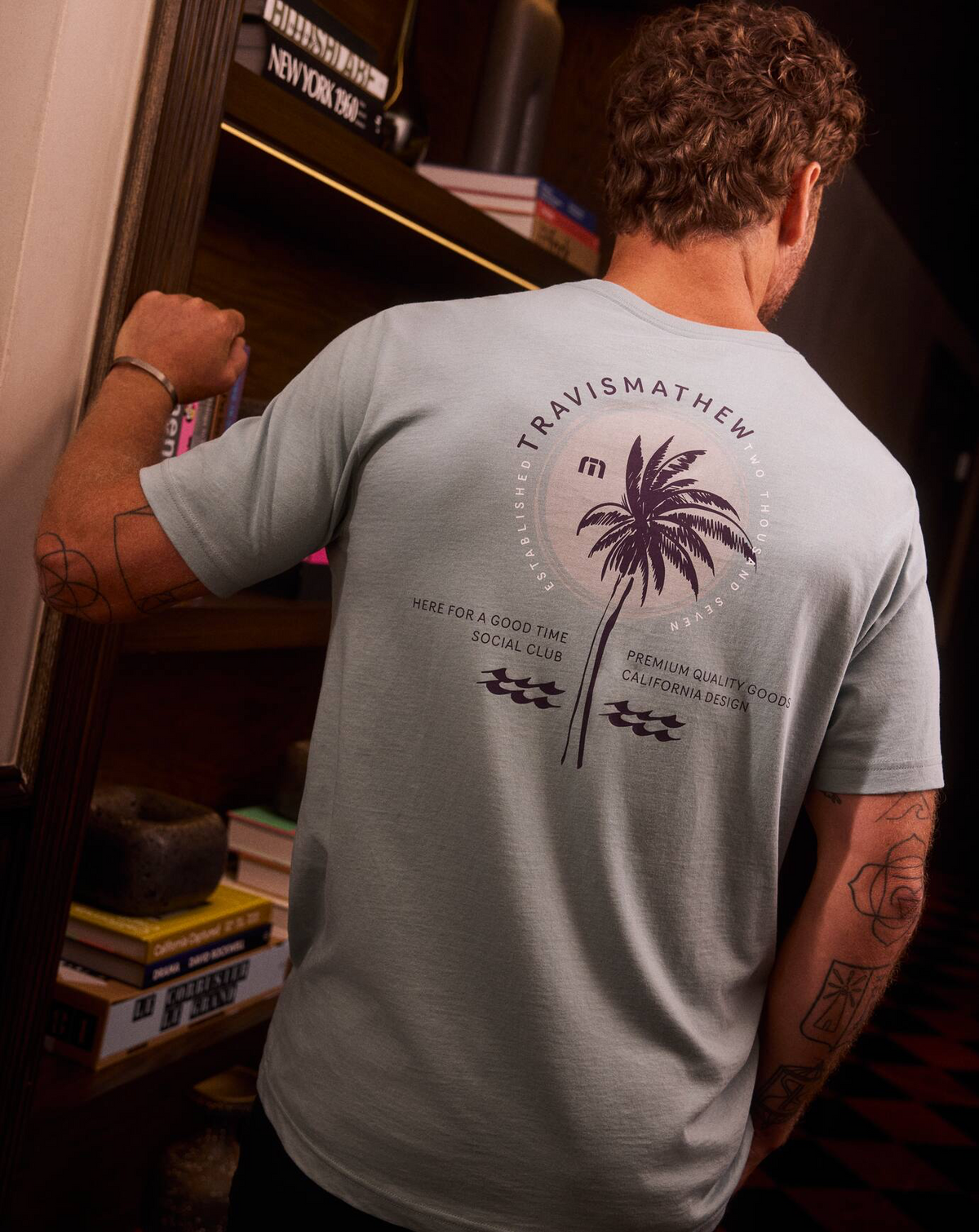 Lifestyle back view of TravisMathew Hold My Calls Tee showing palm graphic, worn indoors