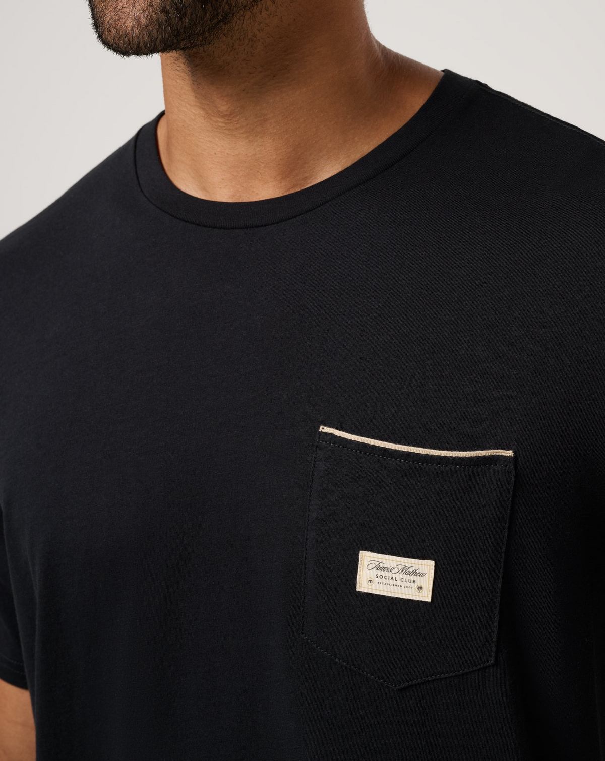 Close-up of TravisMathew Social Club Tee chest pocket detail in black
