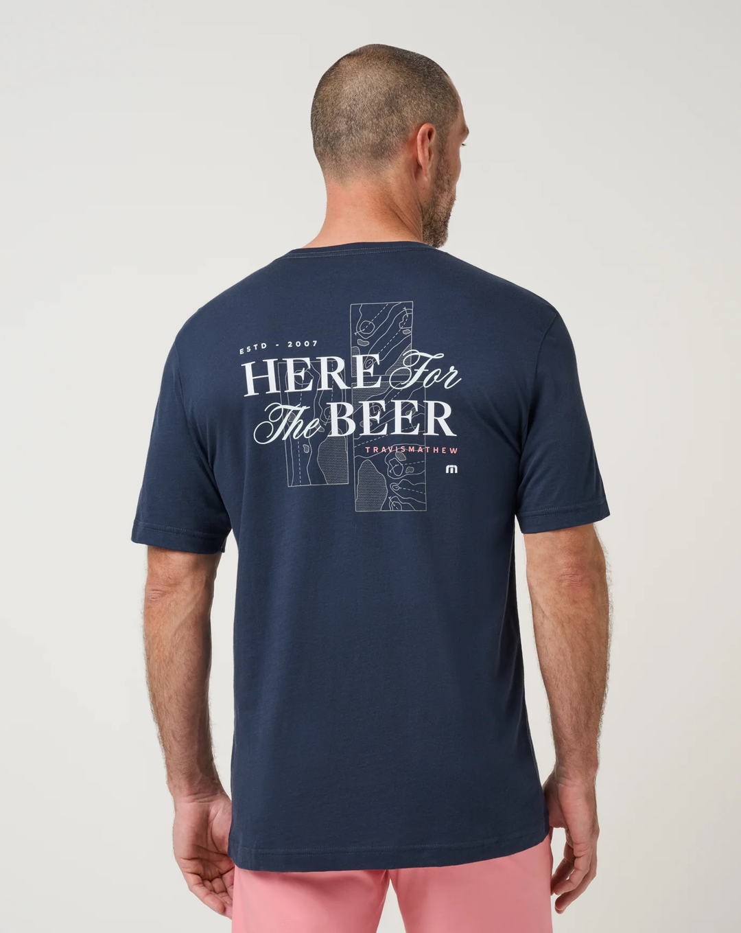 TravisMathew Stout Standard Tee in navy, back view on model showing the Here For The Beer graphic over an aerial golf course sketch