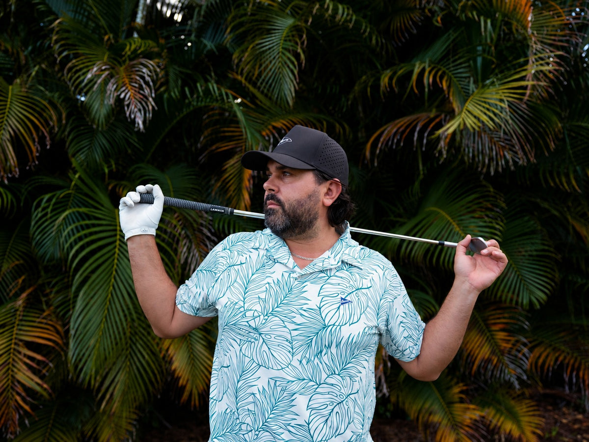Golfer in a white and teal palm print polo holding a club across his shoulders in front of palm trees